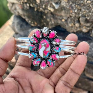 Könnte beinhalten: Ein silbernes Armreif-Armband mit einem ovalen Mittelstein und tropfenförmigen Steinen in Rosa, Türkis und Weiß. Das Armband hat drei parallele Bänder. Der Text "Bohojewels25" ist sichtbar.