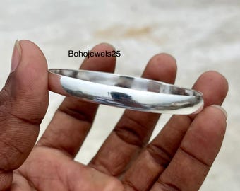 May include: A polished silver bangle bracelet. The smooth, rounded design reflects light, creating a bright, shiny appearance. The bracelet is held in a hand, showcasing its simple, elegant style. The text "Bohojewels25" is visible.