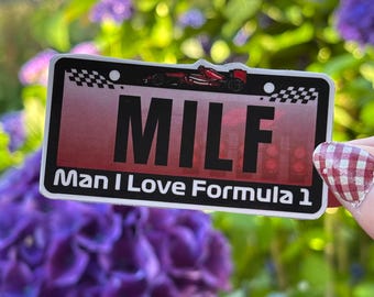 May include: A rectangular sticker with a red and black gradient background. The sticker has the word "MILF" in large, bold, black letters. Below, it reads "Man I Love Formula 1". A race car graphic is at the top.
