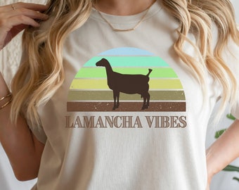 Peut inclure: Un t-shirt beige clair avec une silhouette brune de chèvre devant un graphique de coucher de soleil coloré. Le texte "LAMANCHA VIBES" est imprimé sous le graphique.