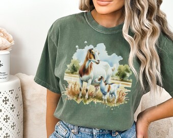 May include: A green t-shirt with a watercolor illustration of two goats in a field. The larger goat is white with brown markings and the smaller goat is brown and white.