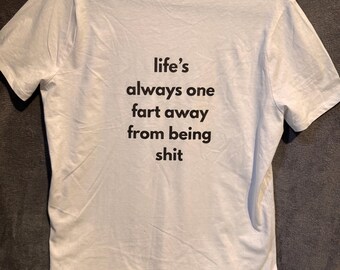 Puede incluir: Camiseta blanca con el texto "life's always one fart away from being shit" en negro. La camiseta está colgada de una percha gris sobre un fondo oscuro. La camiseta tiene cuello redondo y mangas cortas.