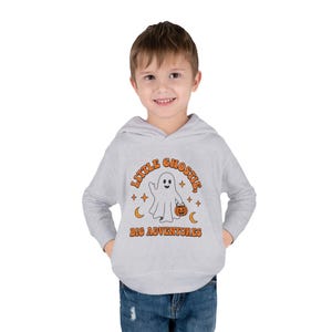 Peut inclure: Un sweat à capuche gris clair avec un motif représentant un fantôme blanc tenant un seau citrouille orange. Le texte "LITTLE GHOSTIE, BIG ADVENTURES" est écrit en orange, entouré de petites étoiles et de croissants de lune orange.