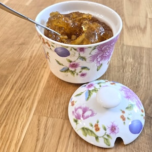 May include: A white ceramic sugar bowl with a floral pattern featuring purple and pink flowers, green leaves, and small orange accents. The bowl is filled with amber-colored marmalade and includes a matching lid and a silver metal spoon.