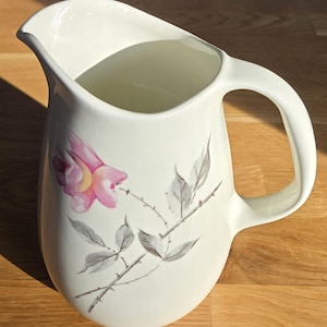 May include: A cream-coloured ceramic jug with a curved handle and a pouring spout. It features a pink and yellow floral design and grey leaves on a thorny branch. This jug is likely used for serving beverages, and is a decorative item.