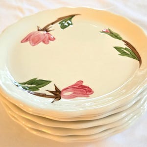 May include: A stack of off-white ceramic plates with scalloped edges. Each plate is decorated with pink floral designs and green leaves. The plates are stacked on top of each other.