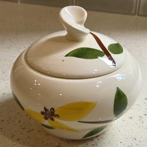 May include: A round, cream-colored ceramic sugar bowl with a matching lid featuring a twisted handle. The surface displays a painted botanical pattern of yellow flowers, green leaves, and brown stems, with a visible fine crackle glaze finish.