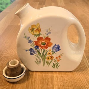 May include: A white ceramic pitcher with a rounded, flat-sided body and a side handle. The front features a colorful floral illustration of yellow, orange, and blue flowers. A separate small white ceramic base with a cork insert is placed nearby.