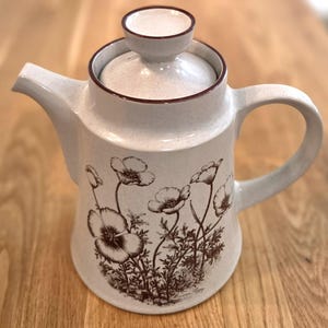 May include: A speckled off-white ceramic teapot with a matching lid featuring a dark brown rim. The body of the teapot is decorated with a detailed dark brown botanical illustration of poppy flowers and foliage, including the text Mexican Poppy.