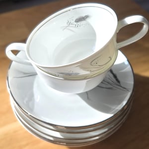 Puede incluir: Juego de taza y platillo blancos con ribete plateado y diseño de trigo. La taza está apilada sobre una pila de platillos. El juego está sobre una superficie de madera.