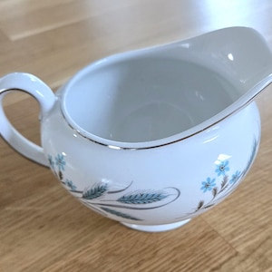 Peut inclure: Crémier en céramique blanche avec une anse et un bec verseur, décoré de motifs floraux bleus et de blé. Le bord est orné d'une bande argentée. Le crémier est posé sur une surface en bois.