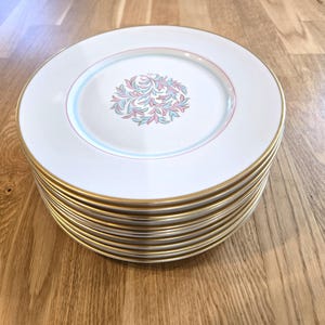 May include: A stack of white ceramic plates with gold rims. Each plate features a central floral design in pink and blue, encircled by a pink and blue ring. The plates are stacked on a wooden surface.