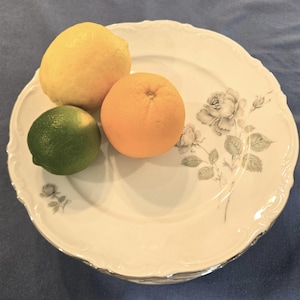 May include: A white porcelain plate with a floral rose design, holding a lemon, an orange, and a lime. The plate has a scalloped edge and a silver trim. The fruits are arranged on the plate.