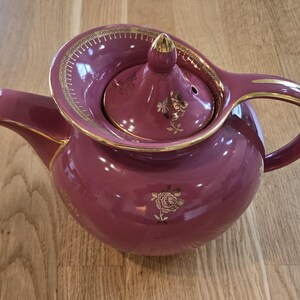 May include: A maroon ceramic teapot with gold detailing and floral patterns. The teapot features a rounded body, a curved handle, and a lid with a gold finial. The spout and rim are also trimmed in gold.