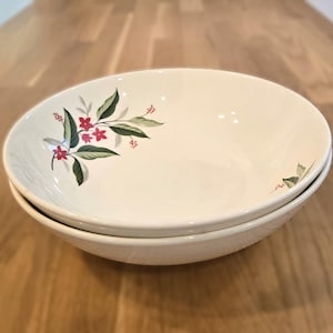 May include: Two white ceramic bowls stacked on top of each other. Each bowl has a floral design with red flowers and green leaves. The bowls are on a light brown wooden surface.