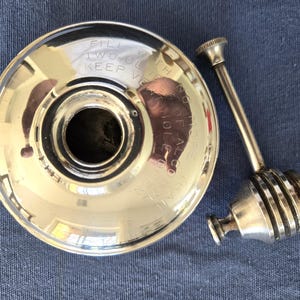 May include: A silver-colored, vintage-style oil lamp with a round base and a separate, attached burner. The base has engraved text that reads "FILL TWO OUNCES KEEP VERTICAL". The burner has a long stem and a decorative top.