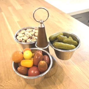 Peut inclure: Un présentoir à trois niveaux avec des bols en acier inoxydable. Les bols contiennent des pistaches, des cornichons et des tomates colorées. Le présentoir a une poignée en bois et repose sur une surface en bois.