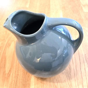 May include: A gray ceramic pitcher with a rounded body and a curved handle. The pitcher has a spout for easy pouring. The glossy finish reflects light, highlighting the smooth surface. This kitchenware item is suitable for serving beverages.