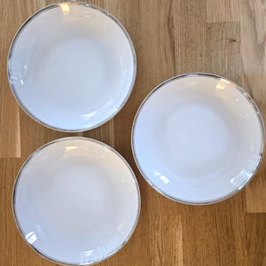 Puede incluir: Tres platos de cerámica blanca con borde plateado dispuestos sobre una superficie de madera. Los platos son redondos con un borde ligeramente curvado. El borde plateado añade un toque de elegancia al diseño.