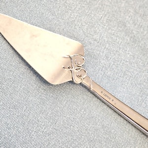 May include: Silver-toned pie server with a triangular blade and a decorative handle. The handle features an ornate design with heart-shaped elements and the word "LENOX" engraved. The server is designed for serving cake or pie.