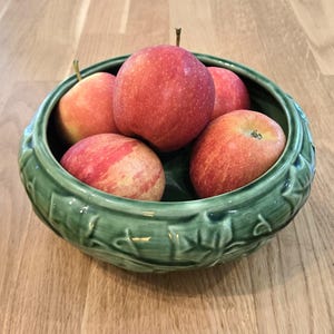 May include: A green ceramic bowl filled with five red and yellow apples. The bowl has a decorative leaf pattern around the rim. The apples are ripe and have a mix of red and yellow coloring.