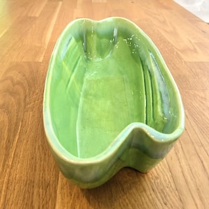 May include: A green ceramic dish with a unique, wavy shape. The dish has a glossy finish and a light green color, with subtle variations in tone. The design features a central, elongated bowl with curved edges and a smooth, reflective surface.