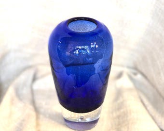 May include: A cobalt blue glass vase with a rounded body and a clear, transparent base. The vase has a small opening at the top and a textured surface. The vase is designed to hold flowers or other decorative items.