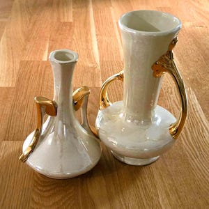 Może przedstawiać: Dwa perłowo-białe wazony ze złotymi uchwytami i zdobieniami. Wazony mają klasyczny kształt, jeden jest krótszy i szerszy od drugiego. Ustawione na drewnianym tle.