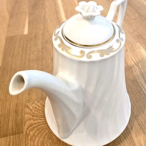 May include: A white porcelain teapot featuring a subtle vertical swirl pattern on the body, a handle, and a spout. The lid is topped with a small white flower-shaped finial and is decorated with a gold and grey scrollwork border design.