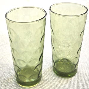 May include: Two green-tinted drinking glasses. The glasses have a textured, bubble-like pattern on the exterior. The glasses are tall and cylindrical with a slightly flared rim. The base of each glass has a decorative design.