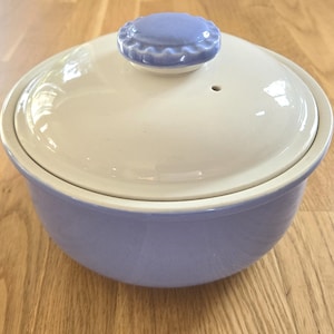 May include: A ceramic casserole dish with a lid. The dish is a pale lavender color, with a cream-colored lid and a light blue knob. The dish is oval-shaped and appears to be unused.