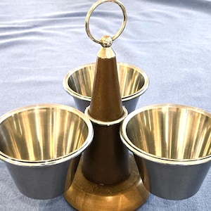 May include: A three-bowl serving stand featuring a central wooden post with a circular metal handle at the top. Three polished stainless steel bowls are attached to the base, arranged symmetrically around the dark brown wooden center column.