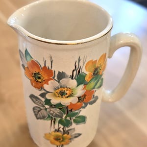 May include: Cream-colored ceramic pitcher with a gold rim and handle. The pitcher is decorated with a floral design featuring orange, white, and yellow flowers with green leaves. A vintage-style decorative piece.