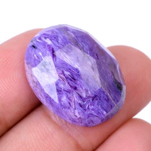 Peut inclure: Une pierre précieuse ovale et facettée, de couleur violette vibrante, avec des motifs tourbillonnants et une surface polie. La pierre est tenue dans une main, mettant en valeur sa taille et ses détails. La couleur varie du lavande clair au violet foncé.