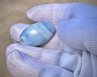May include: A light blue, oval-shaped gemstone with a polished surface and a small hole. The stone has a marbled appearance with white and light blue swirls. The gemstone is held in a light blue fabric.