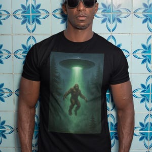 Puede incluir: Camiseta negra con un gráfico de Bigfoot siendo abducido por un OVNI. El diseño incluye un rayo de luz verde, un fondo de bosque y una ilustración detallada del críptido. La camiseta la lleva una persona con gafas de sol.