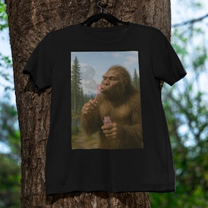 May include: Black t-shirt with a graphic of Bigfoot blowing bubbles. The image shows the creature in a forest setting, holding a bubble wand and a bottle of bubble solution. The shirt is hanging on a black hanger.