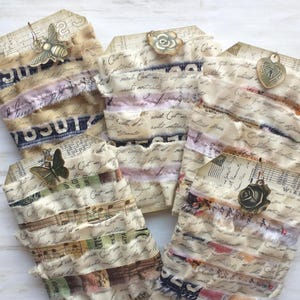 May include: A collection of handmade gift tags with a vintage aesthetic. Each tag features layered fabric strips in various colors and patterns, adorned with metal charms like bees, butterflies, and roses. The tags are made with aged paper and script.
