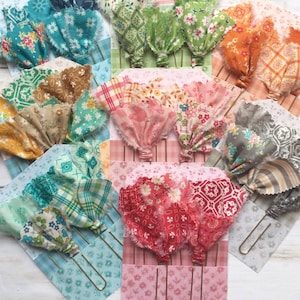 May include: A collection of fabric-topped paper clips featuring various patterned textiles in shades of teal, green, orange, pink, red, and gray. Each clip is attached to a decorative cardstock backing with matching floral, geometric, or plaid prints.