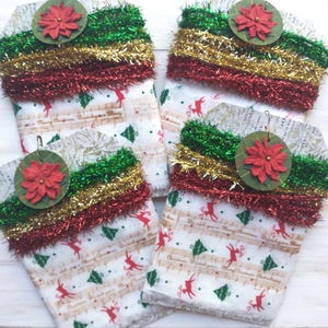 May include: Four rectangular gift tags featuring a white fabric base with a pattern of red reindeer, green Christmas trees, and musical staff lines. Each tag is topped with layers of green, gold, and red tinsel and a circular red poinsettia.