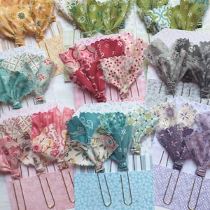 May include: A collection of decorative paper clips featuring fabric tassels in various colors and patterns, including yellow, green, red, pink, blue, gray, and purple. Each fabric piece is tied to a metal paper clip and mounted on patterned cardstock.