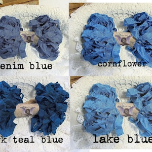 May include: Four bundles of blue fabric ribbon in denim blue, cornflower, dark teal blue, and lake blue. Each bundle is arranged on a white lace surface and labeled with its color name. The ribbons are ruffled and textured.