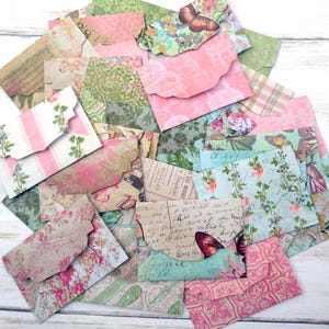 May include: A collection of small, decorative paper envelopes in various patterns and colors. Designs include floral, butterfly, and script motifs in shades of pink, green, and blue. The envelopes have scalloped edges and are arranged on a white wooden surface.