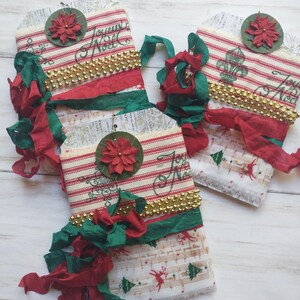 May include: Three rectangular fabric ornaments featuring red and white stripes, gold beaded trim, and green and red ribbon accents. Each ornament is topped with a green circular tag displaying a red poinsettia and the text Joyeux Noel.