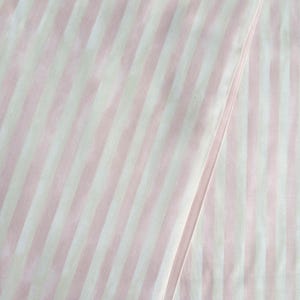 May include: Close-up of a fabric with vertical stripes in alternating shades of pale pink and white. The fabric is folded, creating a diagonal line across the image. The stripes are evenly spaced and run the length of the fabric.