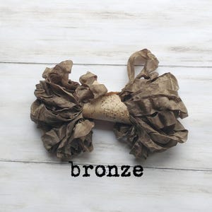 May include: A bronze-colored fabric bow with ruffled edges and a central piece of patterned material. The word "bronze" is printed in black below the bow, which is set against a white wooden background.