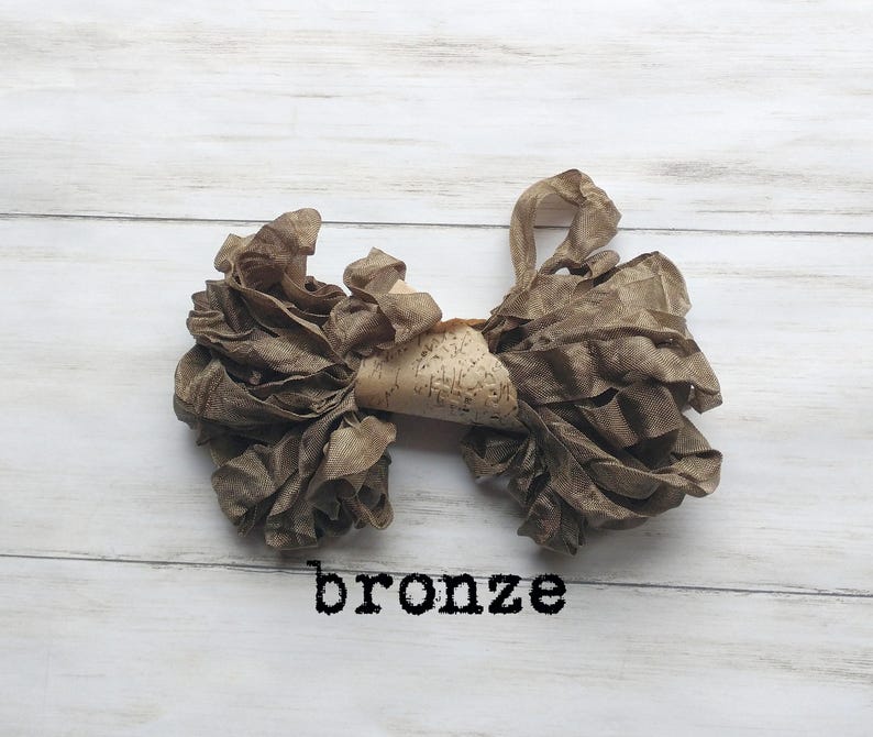 May include: A bronze-colored fabric bow with ruffled edges and a central piece of patterned material. The word "bronze" is printed in black below the bow, which is set against a white wooden background.