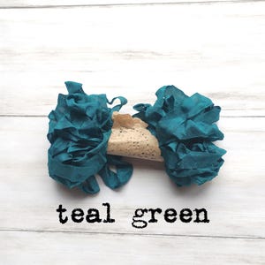May include: Teal green fabric ribbon with a ruffled texture, paired with a beige lace element. The text "teal green" is printed in black below the ribbons. The ribbons are arranged on a white wooden surface.