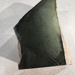 May include: A dark green, irregularly shaped stone with a polished surface. The stone has a triangular form with a rough, beige edge. Fine white lines and small white specks are visible on the surface.