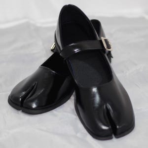 May include: A pair of black Tabi shoes with a split toe design. These shoes have a glossy finish, a Mary Jane strap with a silver buckle, and a low heel. The shoes are designed for comfort and style.